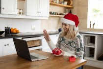 Kaukasische Frau verbringt Zeit zu Hause, sitzt zu Weihnachten mit Weihnachtsmütze in der Küche und benutzt Laptop mit Geschenken auf dem Tisch. Soziale Distanzierung während Covid 19 Coronavirus Quarantäne. — Stockfoto