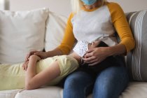 Caucasian woman and her daughter spending time at home together, wearing face masks, sitting on a sofa and embracing. Social distancing during Covid 19 Coronavirus quarantine lockdown. — Stock Photo