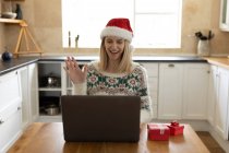 Caucasian woman spending time at home, sitting in kitchen at Christmas wearing Santa hat, using laptop with presents on table. Social distancing during Covid 19 Coronavirus quarantine. — Stock Photo