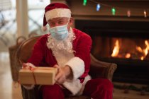 Senior Caucasian man at home dressed as Father Christmas, wearing face mask, sitting on a chair by fireplace, giving presents. Social distancing during Covid 19 Coronavirus quarantine lockdown. — Stock Photo