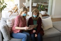 Senior Caucasian woman spending time at home with her adult daughter, sitting on couch, wearing face masks and using tablet computer. Social distancing during Covid 19 Coronavirus quarantine. — Stock Photo