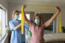 Senior Caucasian woman at home visited by Caucasian female nurse, stretching arms using exercise band, wearing face masks. Medical care at home during Covid 19 Coronavirus quarantine. — Stock Photo