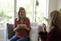 Mujer mayor caucásica pasando tiempo en casa con su hija adulta, sentada en el sofá, bebiendo una taza de té. Distanciamiento social durante la cuarentena del Coronavirus Covid 19. - foto de stock