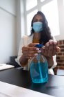 Close up de mulher de raça mista trabalhando em um escritório casual, usando desinfetante e usando máscara facial. Distanciamento social no local de trabalho durante a pandemia do Coronavirus Covid 19. — Fotografia de Stock