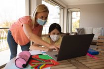 Белая женщина и ее дочь проводят время дома в масках для лица, используя ноутбук во время урока онлайн-школы. Социальное дистанцирование во время изоляции коронавируса Covid 19. — стоковое фото