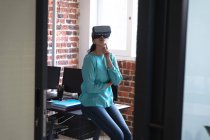 Frau mit gemischter Rasse, die in einem lässigen Büro arbeitet, ihr Headset trägt und auf den virtuellen Bildschirm blickt. Soziale Distanzierung am Arbeitsplatz während der Coronavirus Covid 19 Pandemie. — Stockfoto