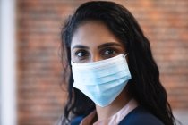 Portrait of mixed race woman working in a casual office, wearing face mask and looking at camera. Social distancing in the workplace during Coronavirus Covid 19 pandemic. — Stock Photo