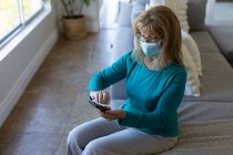 Mulher caucasiana sênior passando tempo em casa, sentada em sua sala de estar limpando smartphone com um lenço. Distanciamento social durante o bloqueio de quarentena do Covid 19 Coronavirus. — Fotografia de Stock