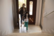 Mujer caucásica y su hija entrando en casa, usando mascarillas, abriendo la puerta. Distanciamiento social durante el bloqueo de cuarentena del Coronavirus Covid 19. - foto de stock