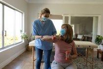 Старша кавказька жінка вдома, яку відвідала кавказька медсестра, розтягнула руку, одягнена в маски для обличчя. Медична допомога вдома під час карантину Covid 19 Coronavirus. — стокове фото