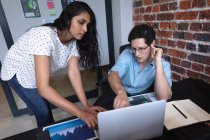 Mujer de raza mixta y hombre caucásico trabajando en una oficina informal, usando una computadora portátil y discutiendo su trabajo. Profesionales creativos de negocios que trabajan en una oficina moderna ocupada. - foto de stock