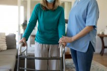 Senior Caucasian woman at home visited by Caucasian female nurse, walking using a walker, wearing face mask. Medical care at home during Covid 19 Coronavirus quarantine. — Stock Photo