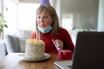 Старша кавказька жінка проводить час удома, сидячи в своїй вітальні з тортом на день народження, одягнувши маску обличчя і використовуючи ноутбук. Соціальна дистанція в Ковиді 19 Коронавірус карантин. — стокове фото
