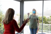 Senior Caucasian woman and her adult daughter at home, wearing face masks and greeting each other through window. Social distancing, health and hygiene during Covid 19 Coronavirus pandemic. — Stock Photo