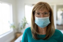 Portrait of a senior Caucasian woman spending time at home, standing in her living room wearing a face mask. Social distancing during Covid 19 Coronavirus quarantine lockdown. — Stock Photo