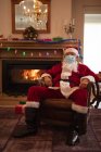 Senior Caucasian man at home dressed as Father Christmas, wearing face mask, sitting on a chair by fireplace. Social distancing during Covid 19 Coronavirus quarantine lockdown. — Stock Photo