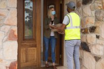 Caucasian woman spending time at home, wearing face mask, receiving a package from delivery man and paying by smartphone. Social distancing during Covid 19 Coronavirus quarantine lockdown. — Stock Photo