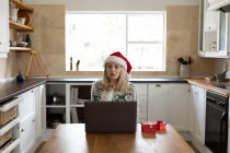 Mujer caucásica pasar tiempo en casa, sentado en la cocina en Navidad con sombrero de Santa, el uso de ordenador portátil con regalos en la mesa. Distanciamiento social durante la cuarentena del Coronavirus Covid 19. - foto de stock