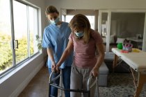 Mujer mayor caucásica en casa visitada por una enfermera caucásica, caminando usando un andador, usando máscaras faciales. Atención médica en el hogar durante la cuarentena del Coronavirus de Covid 19. - foto de stock