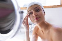 Белая женщина проводит время дома, в ванной, глядя в зеркало, очищая лицо хлопковой салфеткой. Социальное дистанцирование во время изоляции коронавируса Covid 19. — стоковое фото
