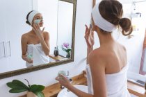 Белая женщина проводит время дома, стоит в ванной, смотрит в зеркало и надевает маску. Социальное дистанцирование во время изоляции коронавируса Covid 19. — стоковое фото