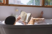 Белая женщина проводит время дома, в ванной, лежит в ванной, читает книгу. Социальное дистанцирование во время изоляции коронавируса Covid 19. — стоковое фото