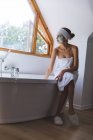 Белая женщина проводит время дома, в ванной комнате в маске, купается сидя на краю ванны. Социальное дистанцирование во время изоляции коронавируса Covid 19. — стоковое фото