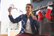 Caucasian woman spending time at home at Christmas, sitting on floor by fireplace in living room, holding ribbon. Social distancing during Covid 19 Coronavirus quarantine lockdown. — Stock Photo