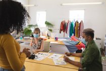 Multiethnische Gruppe von männlichen und weiblichen Modedesignern im Studio, die Gesichtsmasken tragen und über Designs diskutieren. Gesundheit und Hygiene am Arbeitsplatz während der Coronavirus Covid 19 Pandemie. — Stockfoto