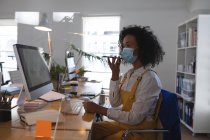 Femme de race mixte travaillant au bureau dans un bureau moderne portant un masque facial et parlant sur un smartphone. Santé et hygiène sur le lieu de travail pendant la pandémie de Coronavirus Covid 19. — Photo de stock