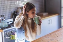 Caucasian woman spending time at home, sitting in kitchen with earphones on, smiling and holding green mug. Social distancing during Covid 19 Coronavirus quarantine lockdown. — Stock Photo