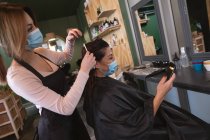 Caucasian female hairdresser working in hair salon wearing face mask, combing hair of female Caucasian customer in face mask. Health and hygiene in workplace during Coronavirus Covid 19 pandemic. — Stock Photo