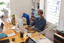 Masculino de raza mixta creativo sentado en el escritorio de una oficina moderna, con una máscara facial y el uso de una computadora. Salud e higiene en el lugar de trabajo durante la pandemia de Coronavirus Covid 19. - foto de stock