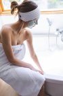 Белая женщина проводит время дома, в ванной комнате в маске, купается сидя на краю ванны. Социальное дистанцирование во время изоляции коронавируса Covid 19. — стоковое фото