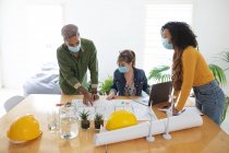 Multi ethnic group of male and female architects in office wearing face masks, discussing over architectural drawing. Health and hygiene in workplace during Coronavirus Covid 19 pandemic. — Stock Photo