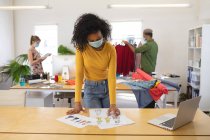 Multiethnische Gruppe von männlichen und weiblichen Modedesignern, die im Atelier arbeiten und Gesichtsmasken tragen und sich distanzieren. Gesundheit und Hygiene am Arbeitsplatz während der Coronavirus Covid 19 Pandemie. — Stockfoto