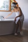 Caucasian woman spending time at home, in bathroom with face mask on, running bath sitting on edge of bathtub. Social distancing during Covid 19 Coronavirus quarantine lockdown. — Stock Photo