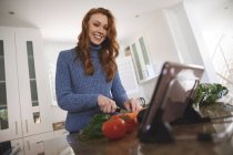 Mujer caucásica pasando tiempo en casa, picando verduras en la cocina, usando su tableta digital, sonriendo. Distanciamiento social durante el bloqueo de cuarentena del Coronavirus Covid 19. - foto de stock