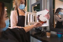 Caucasian female hairdresser working in hair salon wearing face mask, showing hair dyes to female Caucasian customer in face mask. Health and hygiene in workplace during Coronavirus Covid 19 pandemic. — Stock Photo