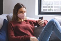 Белая женщина проводит время дома, сидит в гостиной, в маске для лица, используя смартфон. Социальное дистанцирование во время изоляции коронавируса Covid 19. — стоковое фото