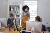 Mixed race and Caucasian female creative business colleague talking in office wearing face masks, male colleague in background. Health and hygiene in workplace during Coronavirus Covid 19 pandemic. — Stock Photo