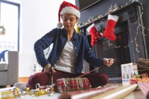 Caucasian woman spending time at home at Christmas, wearing Santa hat, sitting on floor in living room, wrapping present in paper. Social distancing during Covid 19 Coronavirus quarantine lockdown. — Stock Photo