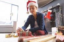 Белая женщина проводит время дома на Рождество в шляпе Санты, сидит на полу в гостиной, заворачивая подарок в бумагу. Социальное дистанцирование во время изоляции коронавируса Covid 19. — стоковое фото