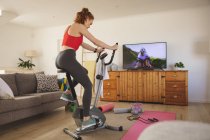 Белая женщина проводит время дома, в гостиной, тренируется на стационарном велосипеде, смотрит телевизор. Социальное дистанцирование во время изоляции коронавируса Covid 19. — стоковое фото