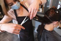 Kaukasische Friseurin, die im Friseursalon arbeitet, trägt Gesichtsmaske, schneidet Haare einer kaukasischen Kundin in Gesichtsmaske. Gesundheit und Hygiene am Arbeitsplatz während der Coronavirus Covid 19 Pandemie. — Stockfoto