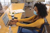 Gemischte Rassenweibchen, die im Rollstuhl am Schreibtisch in einem Büro sitzen, Gesichtsmaske tragen und einen Laptop benutzen. Gesundheit und Hygiene am Arbeitsplatz während der Coronavirus Covid 19 Pandemie. — Stockfoto