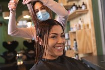 Белая женщина-парикмахер работает в парикмахерской в маске, стрижет волосы белой женщины-клиента. Здоровье и гиперактивность на рабочем месте во время коронавируса Ковид 19 пандемии. — стоковое фото