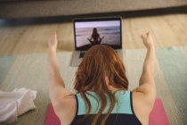 Mujer caucásica pasando tiempo en casa, en la sala de estar, haciendo ejercicio, practicando yoga mientras mira el portátil. Distanciamiento social durante el bloqueo de cuarentena del Coronavirus Covid 19. - foto de stock
