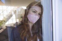 Uma mulher caucasiana atraente, gengibre, passando tempo em casa, na sala de estar, olhando pela janela, usando uma máscara facial. Distanciamento social durante o bloqueio de quarentena do Covid 19 Coronavirus. — Fotografia de Stock