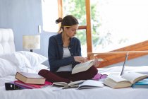 Кавказька жінка проводить час удома, сидячи на ліжку в спальні, навчаючись вдома, тримаючи і читаючи книжки. Соціальна дистанція в Ковиді 19 Коронавірус карантин. — стокове фото